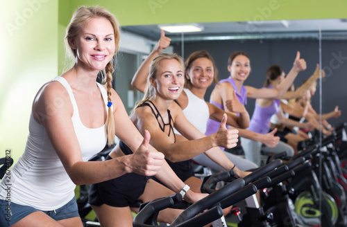Portrait of women on exercise bike gesturing thumbs up at gym