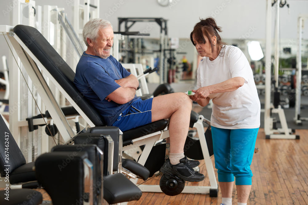 Senior man suffering from knee pain after wokout. Elderly woman applying ointment cream on injured mans leg at gym. Human expression of pain. Senior man suffering from knee pain after wokout. Elderly woman applying ointment cream on injured mans leg at gym. Human expression of pain.