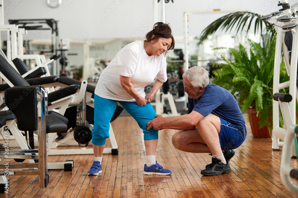 Fototapeta premium Older woman injured leg during fitness training. Man touching injured female knee t gym. Injury from workout concept.