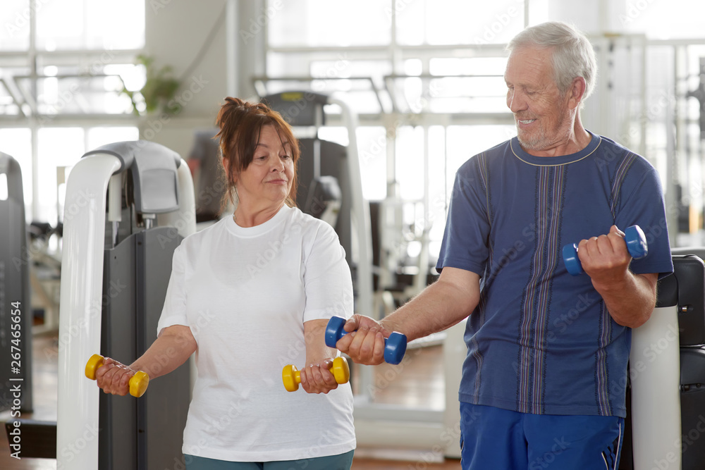 Fototapeta premium Caucasian senior people exercising at gym. Elderly couple lifting dumbbells together at fitness center. People, sport, healthy lifestyle.