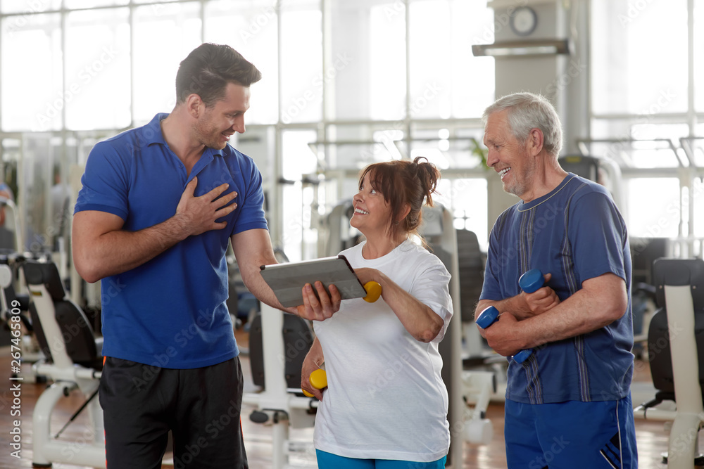 Fototapeta premium Personal male trainer showing something on digital tablet. Happy senior couple at gym. Sport, fitness, technology, lifestyle and people concept.