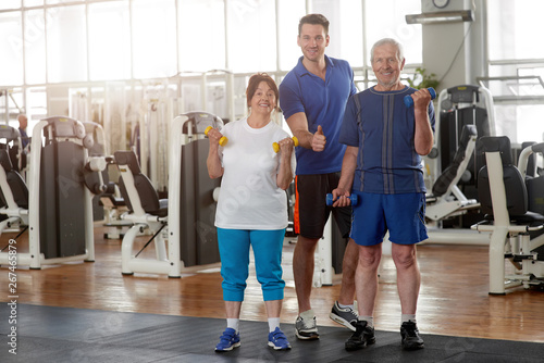 Elderly people with personal trainer at gym. Portrait of fitness trainer with senior couple showing thumb up. Healthy lifestyle for energy and longevity.