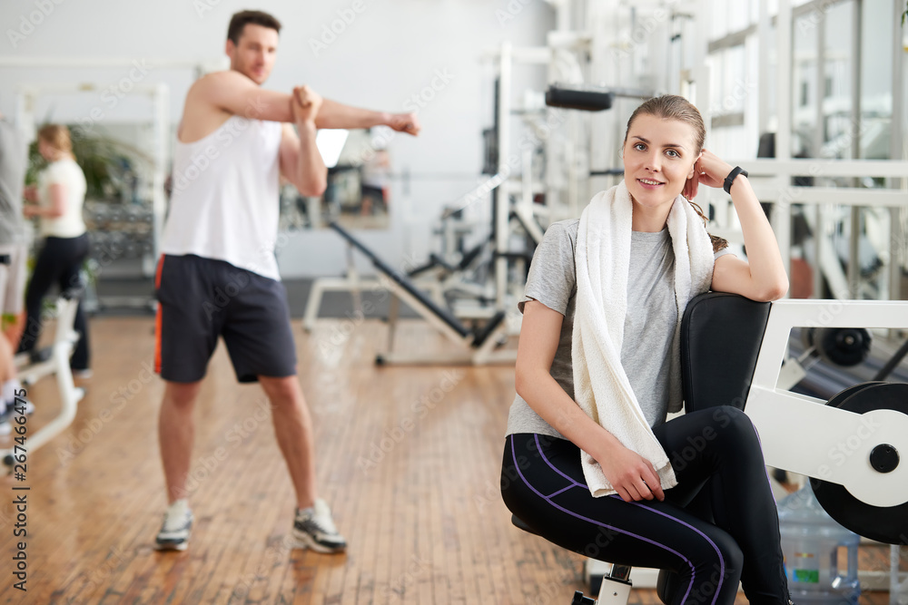 Portrait of tired woman having rest after workout. Beautiful sporty girl with towel and sweat looking at camera at modern gym. Handsome muscualr man working out on the background. Portrait of tired woman having rest after workout. Beautiful sporty girl with towel and sweat looking at camera at modern gym. Handsome muscualr man working out on the background.