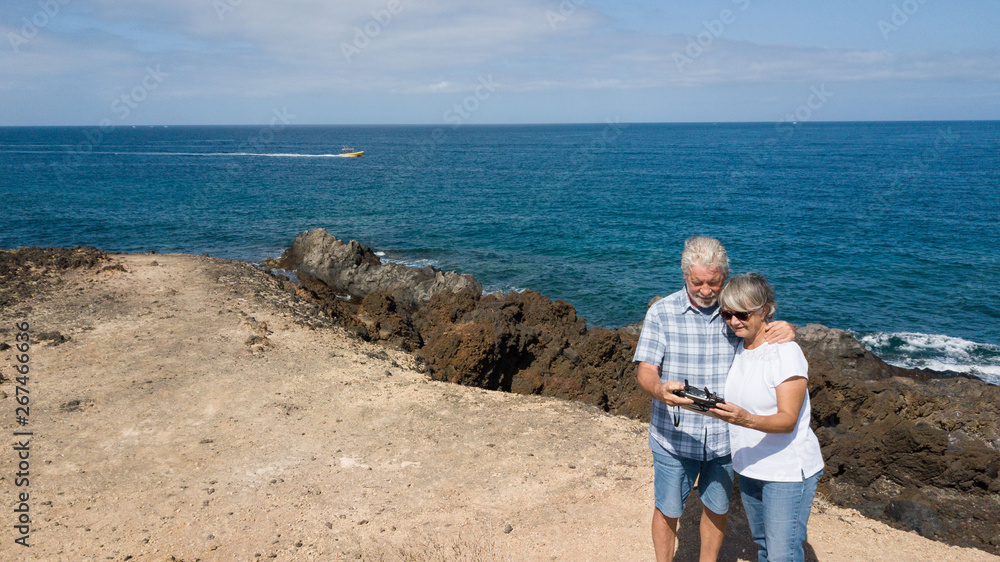 Two people senior looking at the device control of the drone. Blue ocean behind them. Enjoying the excursion and relaxing. Beautiful background of sky and sea