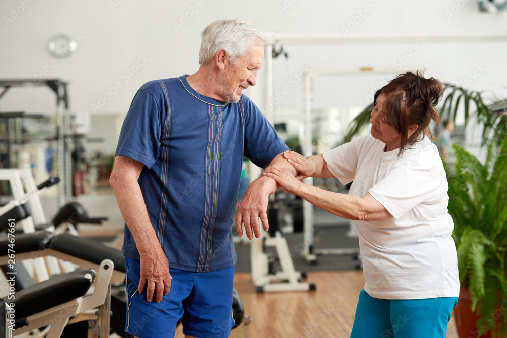 Senior man feeling pain in elbow during workout. Elderly woman holding injured arm of man at gym. Human expression of pain. Senior man feeling pain in elbow during workout. Elderly woman holding injured arm of man at gym. Human expression of pain.