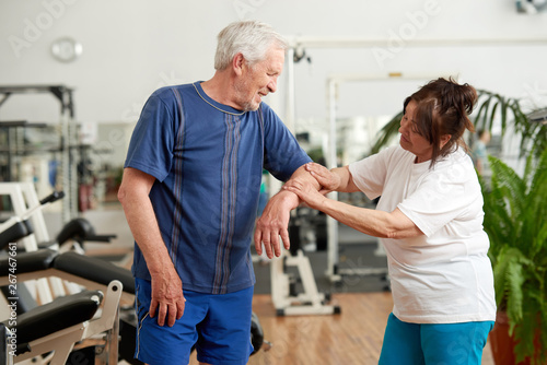 Senior man feeling pain in elbow during workout. Elderly woman holding injured arm of man at gym. Human expression of pain.