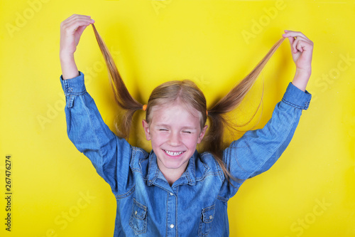 Laughing funny little girl standing on yellow background and holding ponytails