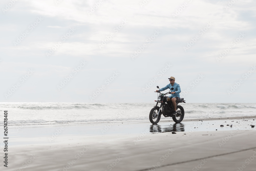 Obraz premium Man in cap riding motorcycle on beach. Moto cross dirtbiker on beach sunset on Bali. Young hipster male enjoying freedom and active lifestyle, having fun on a bikers tour.