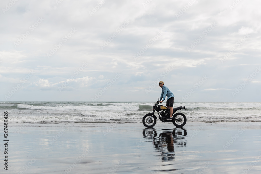 Fototapeta premium Man in cap riding motorcycle on beach. Moto cross dirtbiker on beach sunset on Bali. Young hipster male enjoying freedom and active lifestyle, having fun on a bikers tour.