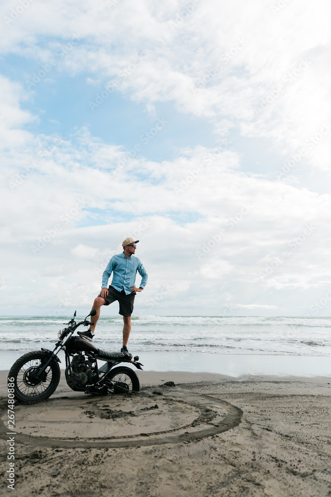 Fototapeta premium Man stands on a motorcycle. Ocean beach Bali. back wheel of a motorcycle in sand. male person looking for inspiration while standing on sportive motorbike. Young man surfer enjoying recreation near to