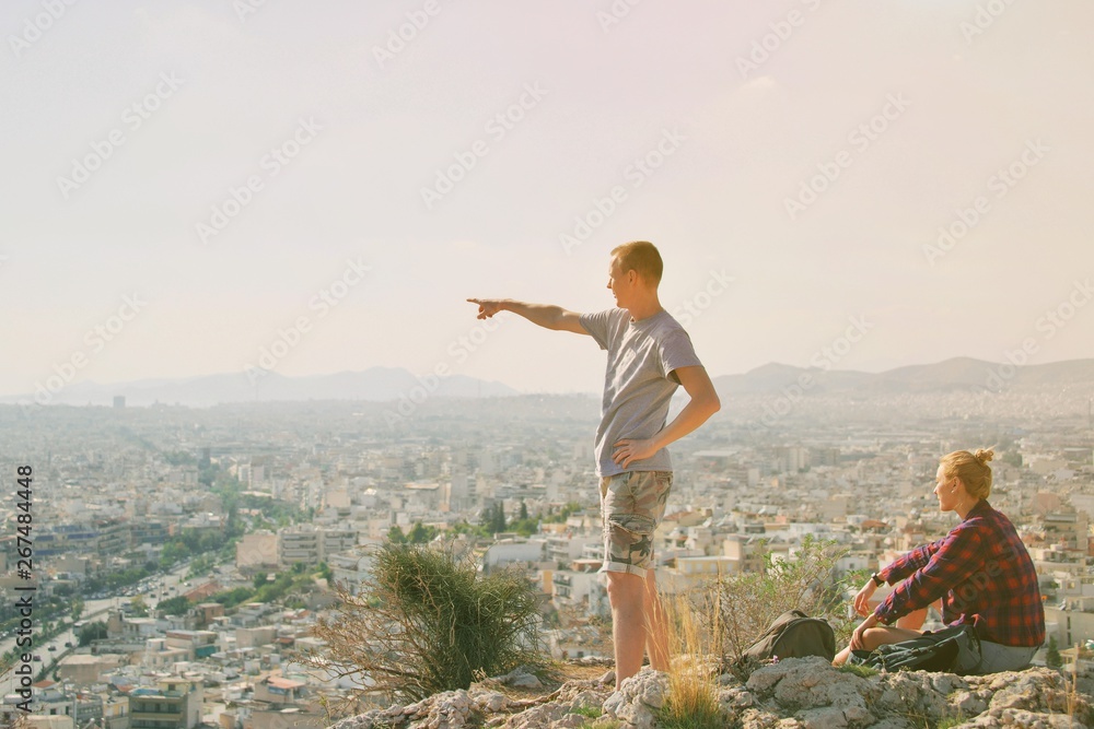 Couple Man and Woman on top of mountain enjoying beautiful landscape cityscape Athens Greece on background. Tourists relax on hill Athens in summer. Famous Athens city in Europe. traveling adventure.