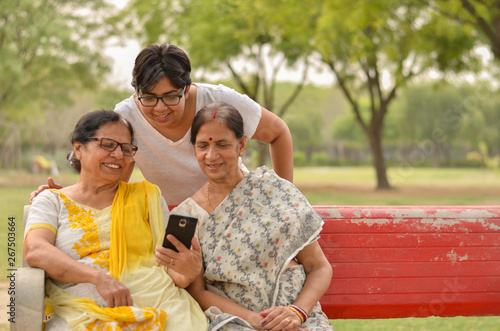 Wallpaper Mural A young Indian girl with two senior women looking at the mobile phone screen while sitting on a red park bench in New Delhi, India. Concept Mother's day Torontodigital.ca