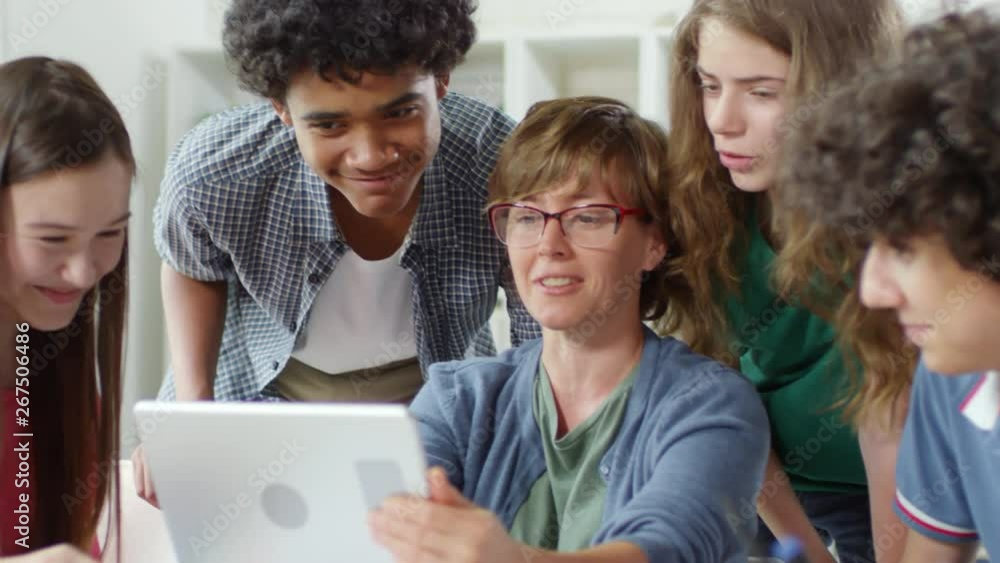 Medium shot of teenagers gathered around their middle-aged female teacher sitting at table with tablet in her hands and showing them interesting information Medium shot of teenagers gathered around their middle-aged female teacher sitting at table with tablet in her hands and showing them interesting information