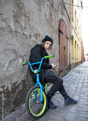 European teenager with dreadlocks in black hat sits on a bike in the old town and looks into his phone