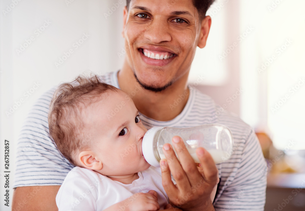A portrait of father bottle feeding a small toddler son indoors at home. A portrait of father bottle feeding a small toddler son indoors at home.