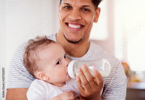 A portrait of father bottle feeding a small toddler son indoors at home.