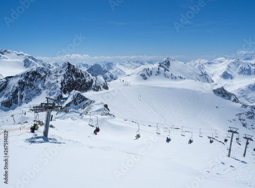 Scenic view from top of Wildspitz on winter landscape with snow covered mountain slopes and pistes and skiers on chair lift at Stubai Gletscher ski resort at spring sunny day. Blue sky background