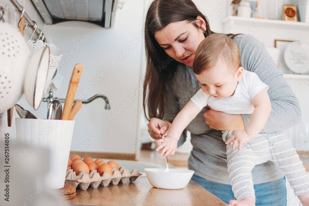 Young mother brunette woman with baby boy in arms cooking food in white modern kitchen at home