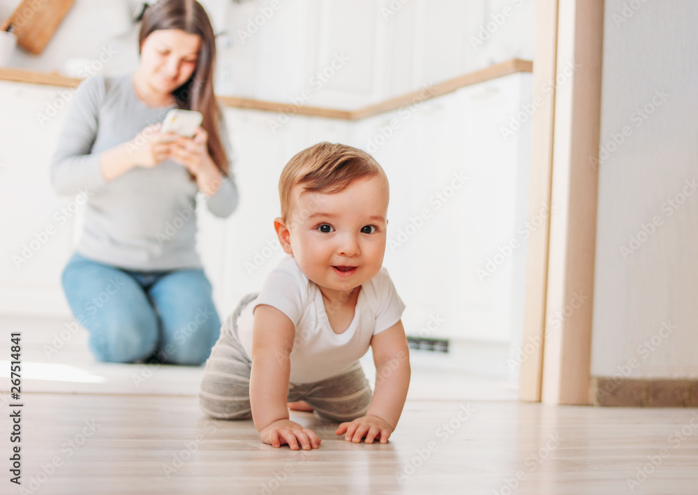 The cute baby boy is exploring the world, while mother surfing in your smartphone in kitchen The cute baby boy is exploring the world, while mother surfing in your smartphone in kitchen