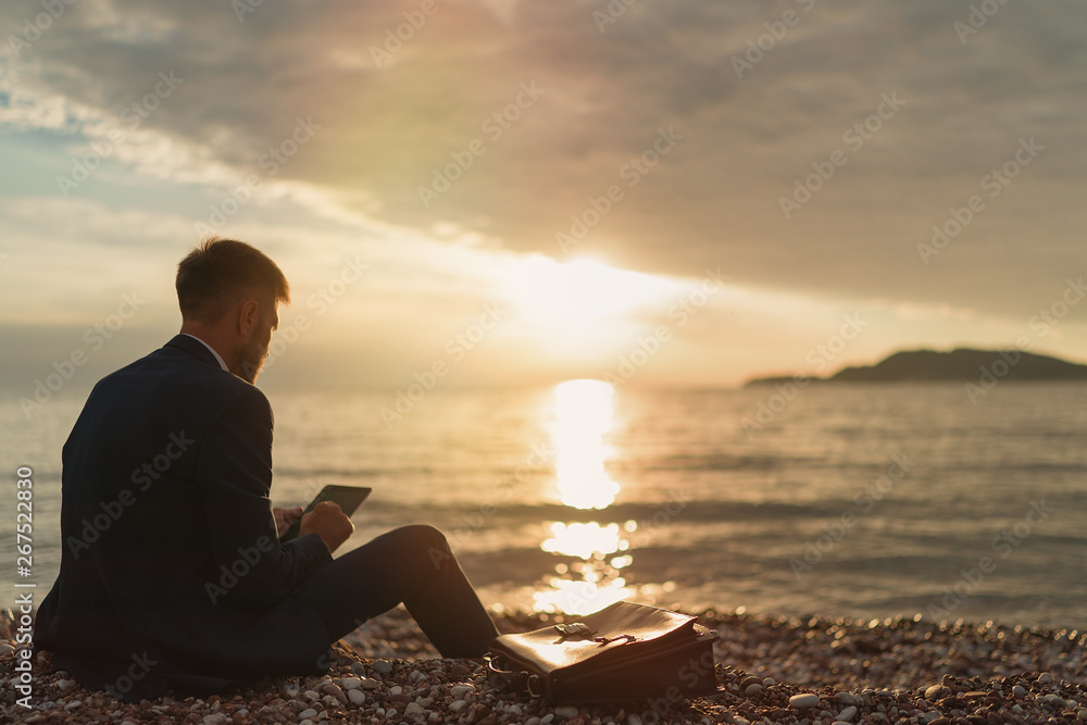 Businessman in a suit using tablet while sitting on the sand of the beach during sunset