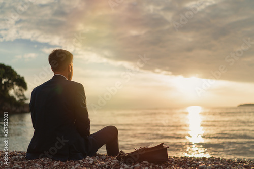 Businessman sitting on the sand of the beach during sunset