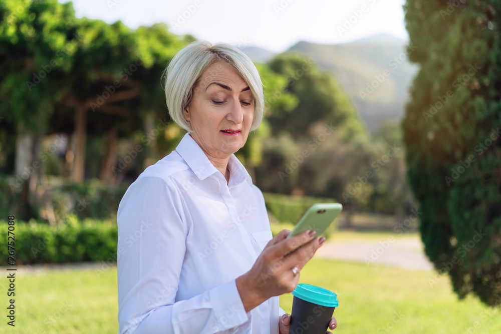 Mature woman using on mobile phone while resting at the park. Senior lady holding takeaway coffee Mature woman using on mobile phone while resting at the park. Senior lady holding takeaway coffee