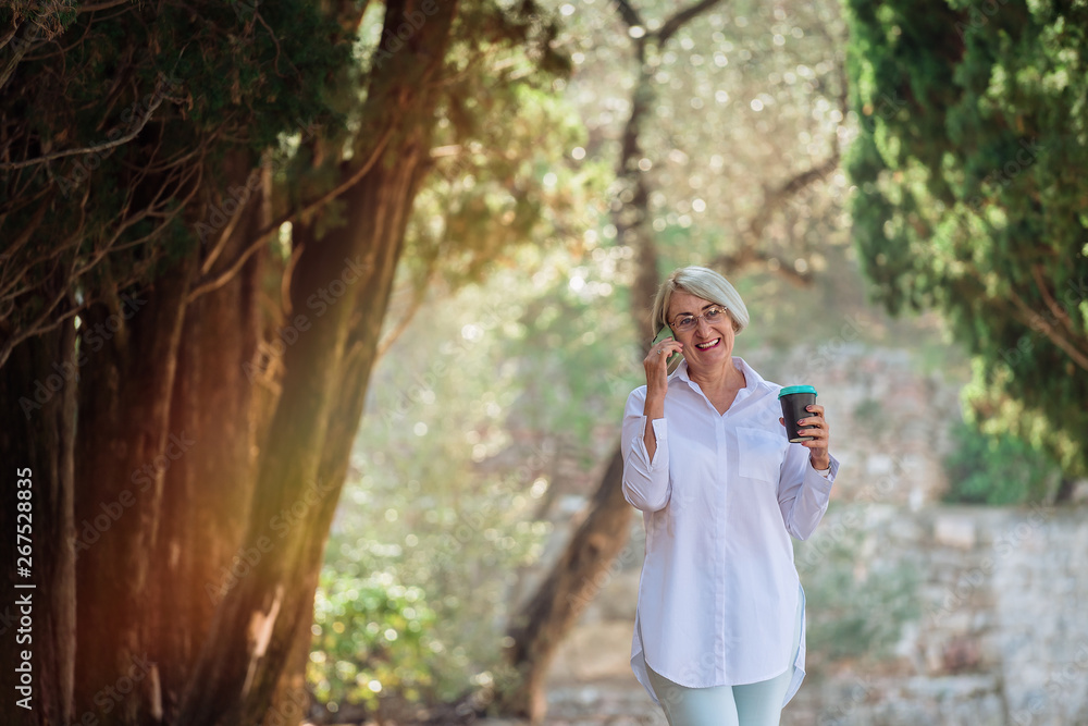 Mature woman talking on mobile phone while resting at the park. Senior lady holding takeaway coffee Mature woman talking on mobile phone while resting at the park. Senior lady holding takeaway coffee