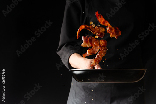 Obraz na plátně The chef prepares bacon, roasting slices of bacon on a black background, horizon