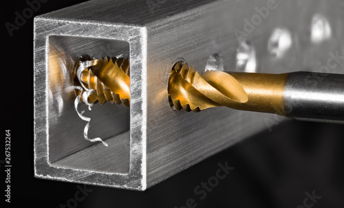 Tapping tool. Steel tap drill coated by titanium. Spiral swarf. Cutting of thread in hole. Aluminum alloy profile. Working cutter. Abstract chip machining detail on black background. Metal production.