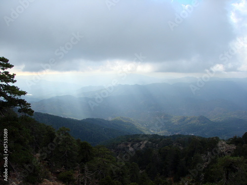 An unusual natural picture of the rays of the sun that penetrates through a heavy gray cloud to dense forests on the slopes of the mountains. © Hennadii