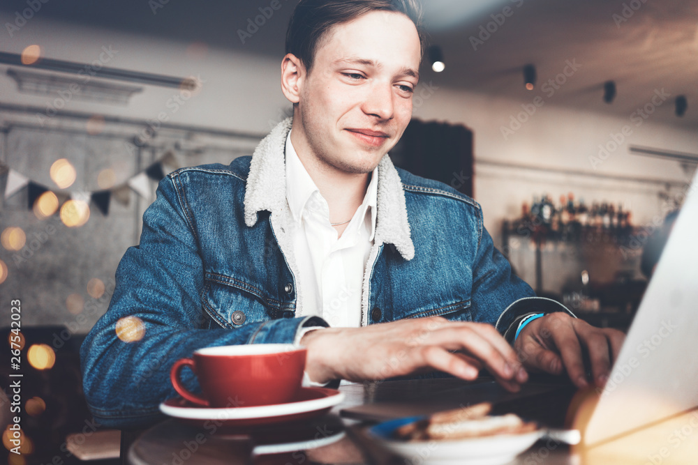Fototapeta premium Young smiling man typing message on laptop keyboard. Sitting in city cafe in smart casual dress