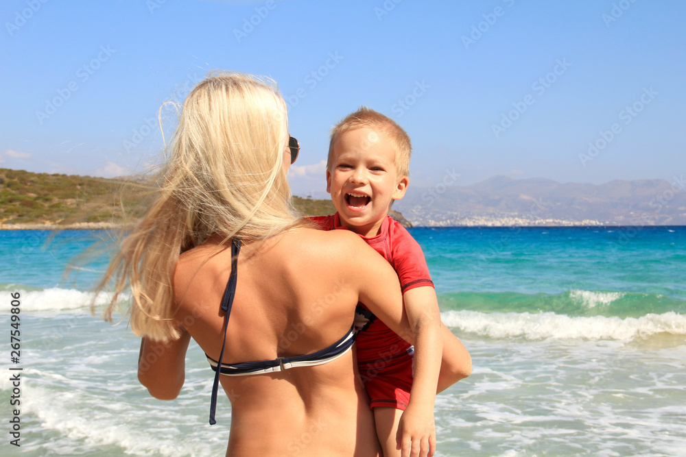 Young slim woman holding her son on the beach by the clear blue sea. Little boy in UV filter clothes is smiling, mother and her cute child are having fun, holiday with children concept