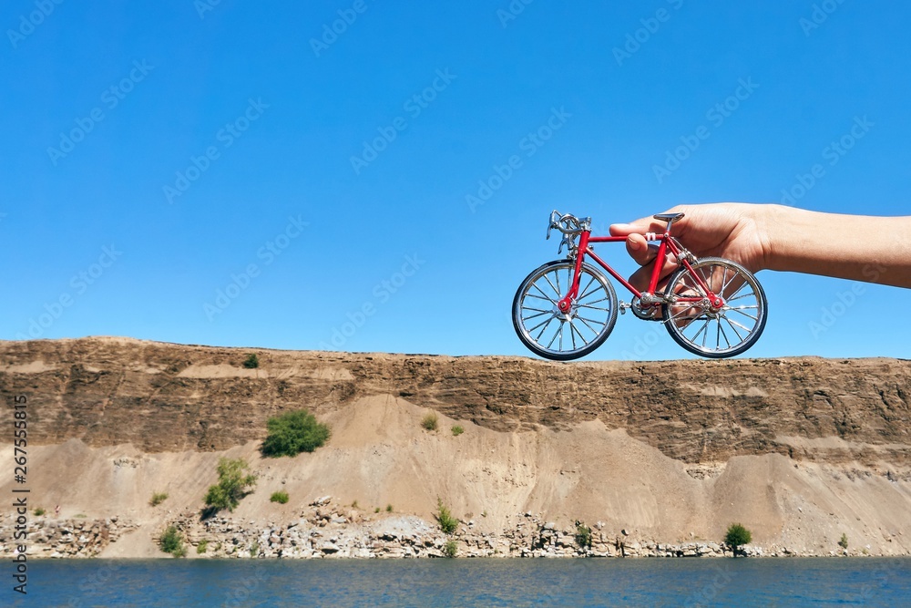 red metal toy bike in the hands against the background of a cliff in the distance with an optical illusion of a big bike on a clear sunny day in summer while traveling