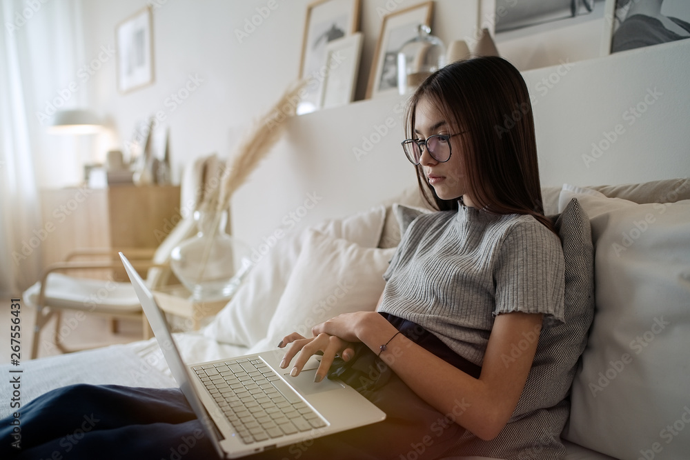 Cute teenager girl doing homework lying on bed at home. Young pretty girl wearing glasses studying online with laptop, distance learning, self education Cute teenager girl doing homework lying on bed at home. Young pretty girl wearing glasses studying online with laptop, distance learning, self education