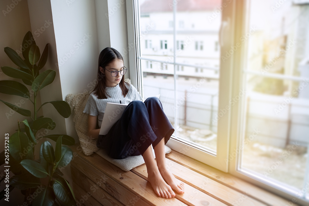 Cute teenager girl reading book at home while sitting on window sill Cute teenager girl reading book at home while sitting on window sill