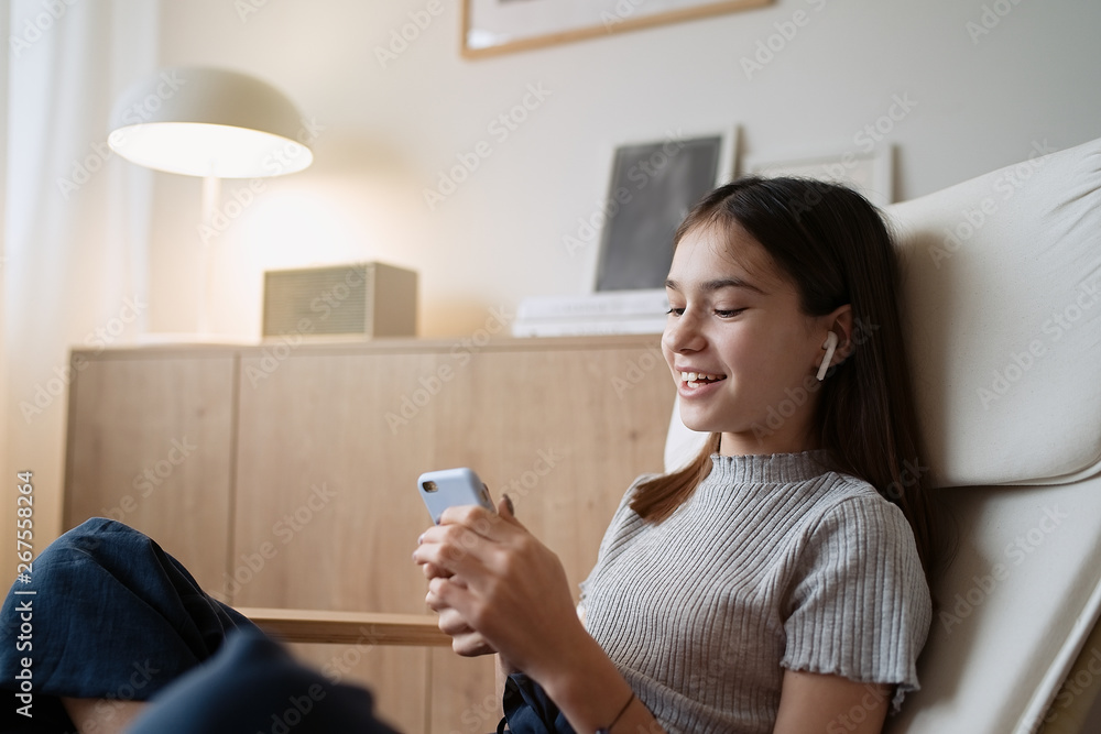Cute teenage girl sitting on armchair at home and using mobile phone with headphones        