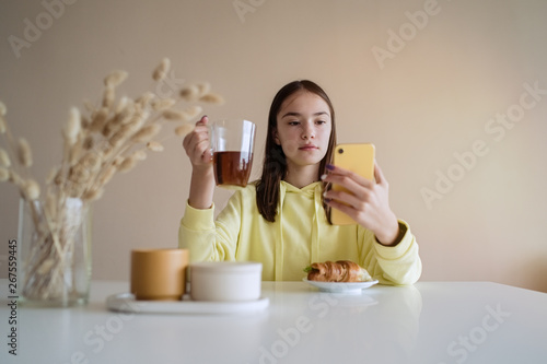 Cute teen girl eating breakfast in kitchen while using on her smart phone 