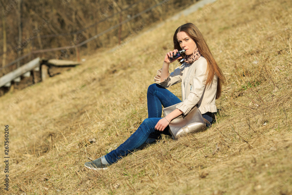 Vape teenager. Young pretty white girl in casual clothing smoking an electronic cigarette on the hillside on a sunny day in the spring. Bad habit. Vaping activity.