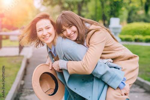 Canvas Print Two adult playfull good looking women sisters in trendy coat piggybacking her pretty girlfriend in spring park outdoor