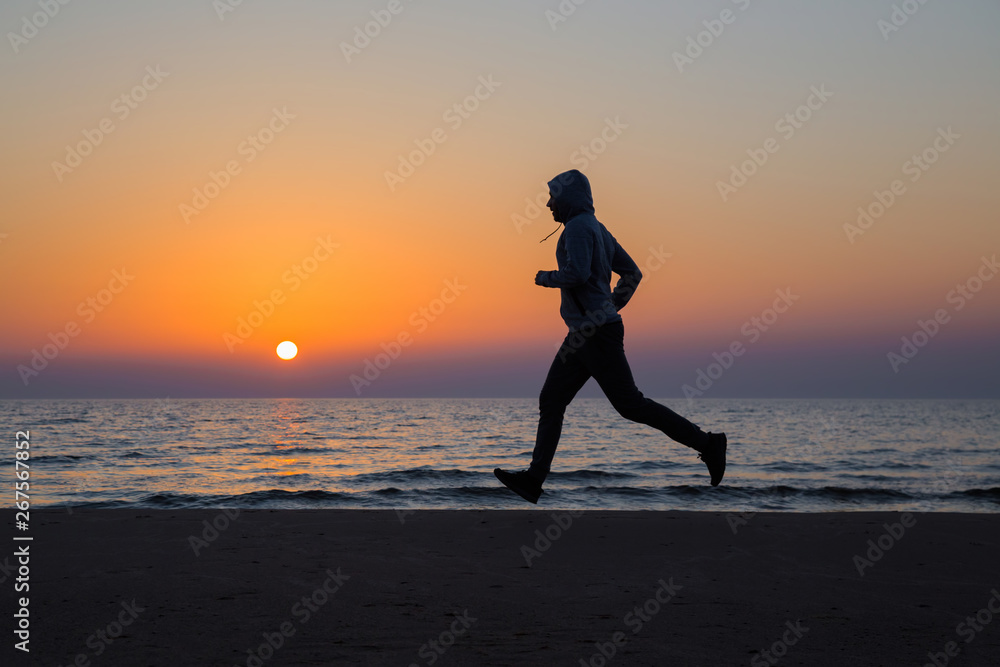 Alone man running on sand beach. Beautiful colorful sunset sky. Sportsman in tracksuit with hood. Enjoying sport in evening. Outdoor workout. Daily active lifestyle. Side view. 