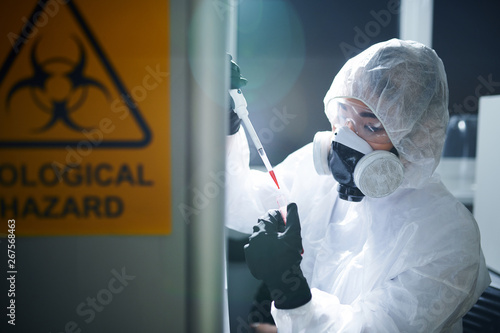 Concentrated laboratory scientist in protective workwear, rubber gloves, goggles and respirator complying with safety while working with infected substance photo