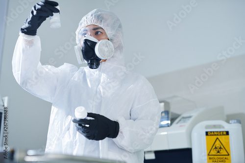 Serious thoughtful toxic laboratory worker in biohazard suit and respirator standing in isolation room and checking bottles of substances while studying drugs for treatment in laboratory