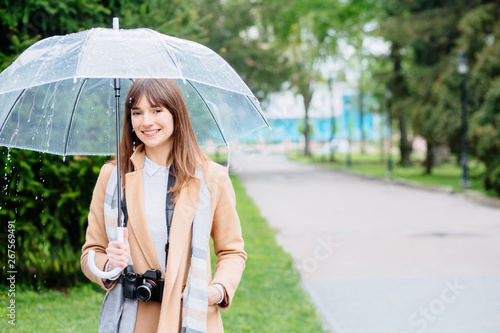 Spring happy brunette woman in beige coat with camera transparent umbrella in spring park on green outdoors background. Life style, travel concept.Model under rain looking up.