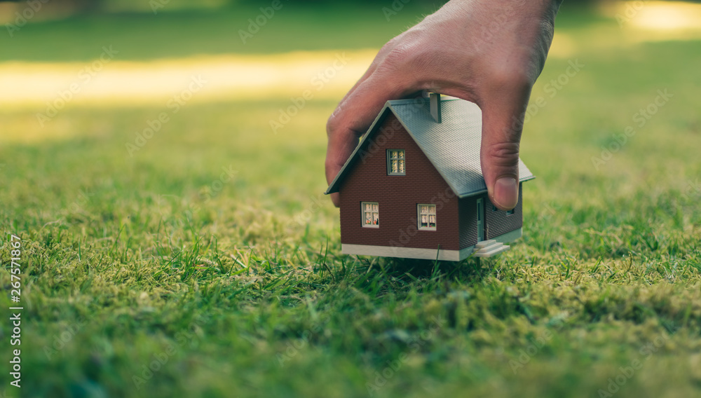 Concept of selling a house. A hand is holding a model house above green meadow. Concept of selling a house. A hand is holding a model house above green meadow.