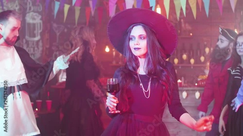 Attractive woman disguised as a witch dances with a glass of blood at a halloween party. Friends gathered at a halloween party in scary costumes.