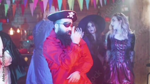 Bearded man in pirate costume dances at a halloween party. People in different scary costemes dancing in the background in a decorated pub.