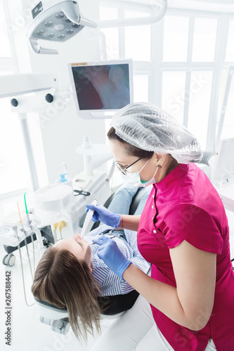 A professional dentist woman in glasses and overalls examines the oral cavity of a young girl in the dental chair using an intraoral stamotological video camera with LED illumination