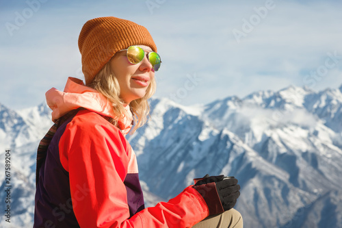 Close-up Portrait of a beautiful girl in a hat and sunglasses with a mug of coffee or tea while sitting on a stone in the mountains. The concept of tourism and recreation in the mountains