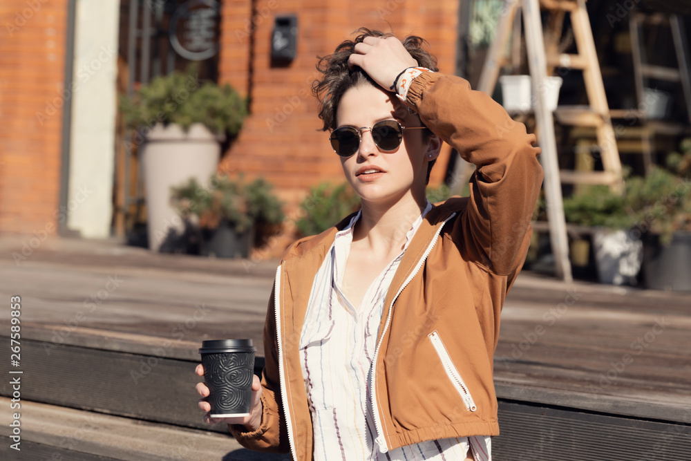 Young beautiful girl with a stylish haircut sits on the street and drinks coffee.