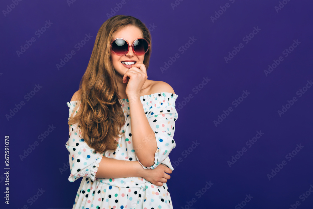 Young smiling woman Photo with copy space Long-haired girl in polka dot dress and big pink sunglasses is smiling against a purple background Young smiling woman Photo with copy space Long-haired girl in polka dot dress and big pink sunglasses is smiling against a purple background